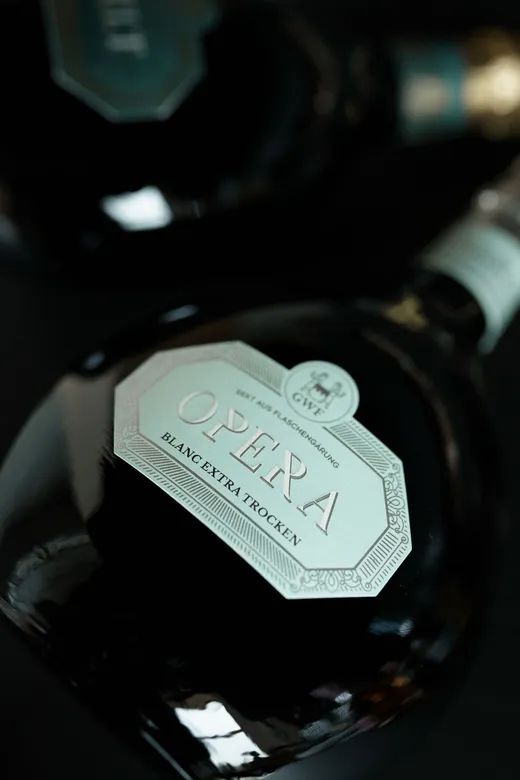 Nahaufnahme von zwei liegenden Weinflaschen aus dunklem Glas, wobei das Etikett „Opera Blanc Extra Trocken“ auf der vorderen Flasche deutlich zu erkennen ist. Der Hintergrund ist unscharf und dunkel.