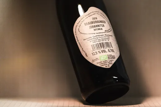 Eine Nahaufnahme einer auf der Seite liegenden Weinflasche, auf deren Etikett folgende Angaben zu sehen sind: „2016 Grauburgunder Johanniter, Gutswein, 12,5 % vol, 0,75 l“. Auf dem Etikett sind außerdem Zertifizierungslogos und deutscher Text zu sehen.