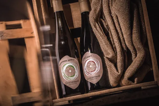 Zwei Flaschen Gustavshof-Wein mit kunstvollen Etiketten stehen in einer Holzkiste neben einem aufgerollten Jutesack und schaffen so eine rustikale und gemütliche Atmosphäre.