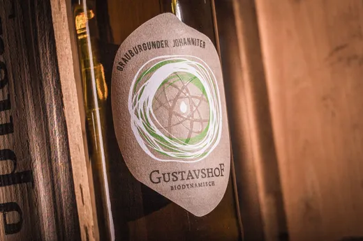 Nahaufnahme einer Weinflasche mit dem Etikett „Gustavshof Biodynamisch, Grauburgunder Johanniter“, auf dem ein grün-weißes kreisförmiges Motiv vor einem rustikalen Holzhintergrund zu sehen ist.