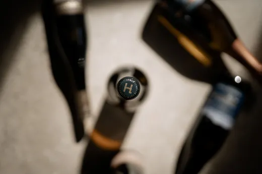 Eine Ansicht von oben auf einen Flaschenverschluss mit dem Buchstaben „H“, umgeben von verschwommenen Weinflaschen auf einer hellen Oberfläche, wobei das Sonnenlicht Schatten wirft.