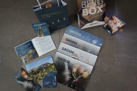 Eine Flat-Lay-Aufnahme von Werbematerialien der Marke Herxheim, darunter Broschüren, Postkarten, ein Faltblatt, Geschenktüten und eine „Berg in a Box“-Weinverpackung, die alle auf einer grauen Fläche angeordnet sind.