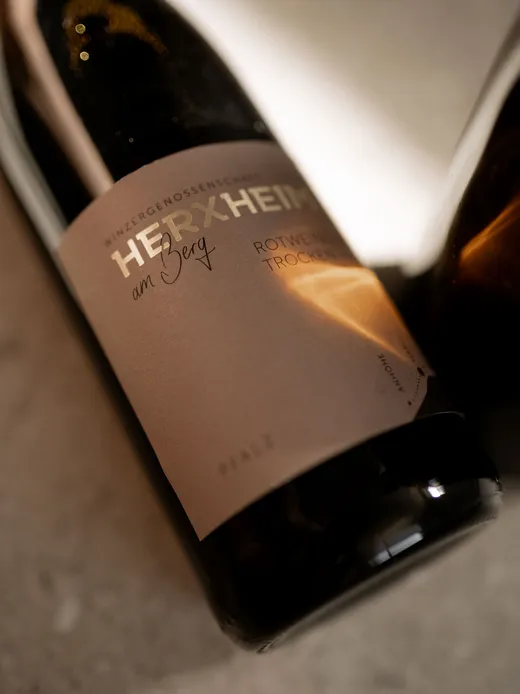 Eine Nahaufnahme einer Weinflasche mit der Aufschrift „HERXHEIM am Berg“, auf deren Etikett ein Schatten und warmes Licht zu sehen sind. Die Flasche steht auf einer hellen Unterlage.