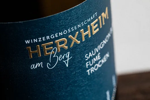 Nahaufnahme eines dunkelblauen Weinetiketts mit gold-weißer Beschriftung, auf der „Winzergenossenschaft HERXHEIM am Berg“ und „Sauvignon Fumé Trocken“ zu lesen sind.