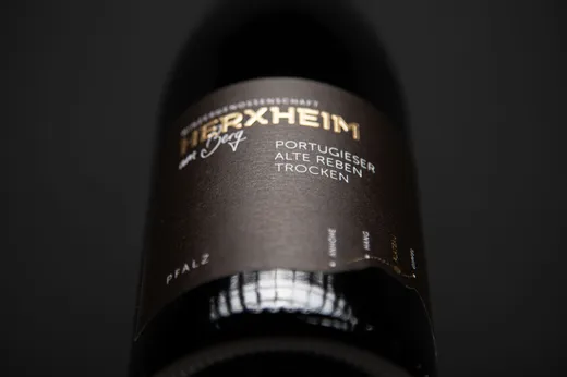 Eine Nahaufnahme einer dunklen Weinflasche mit einem schwarzen Etikett, auf dem „Herxheim Portugieser Alte Reben Trocken“ steht, mit gold-weißer Schrift vor einem unscharfen dunklen Hintergrund.