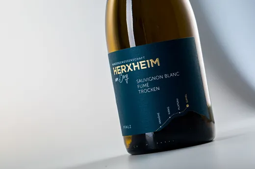 Eine Nahaufnahme einer stehenden Weinflasche mit einem dunkelblaugrünen Etikett, auf dem „Herxheim“ und „Sauvignon Blanc Fumé Trocken“ zu lesen ist. Auf dem Etikett ist außerdem die Region „Pfalz“ angegeben, und es weist minimalistische goldene Akzente auf.