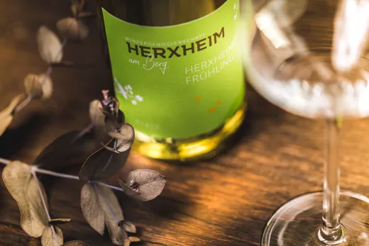 Eine Nahaufnahme einer grünen Weinflasche mit der Aufschrift „HERXHEIM am Berg HERXHEIMER FRÜHLING“ auf einer Holzunterlage, daneben ein Weinglas und einige getrocknete Blätter.