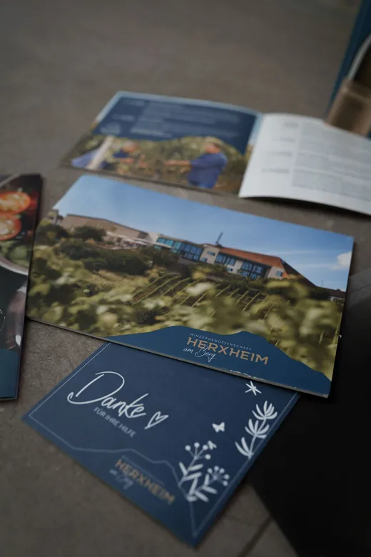 Eine Nahaufnahme mehrerer Broschüren und Karten auf einer Fläche, darunter eine blaue Karte mit der Aufschrift „Danke“ und Herzen sowie eine weitere mit dem Foto eines Gebäudes und dem Text „Herxheim“. Im Hintergrund sind einige unscharfe Prospekte zu sehen.