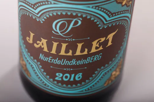Nahaufnahme eines Weinetiketts mit kunstvollen türkisfarbenen und goldenen Motiven. Auf dem Etikett stehen in fetten Buchstaben die Aufschriften „JAILLET“, „NurErdeUndkeinBerg“ und „2016“.