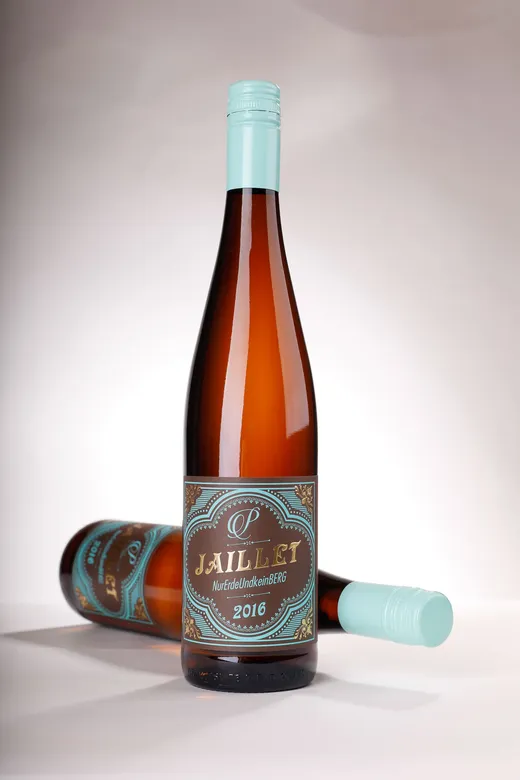 Zwei braune Weinflaschen aus Glas mit türkisfarbenen Verschlüssen und dekorativen Etiketten mit der Aufschrift „Jaillet NurErdeUndkeinBerg 2016“. Eine Flasche steht aufrecht, während die andere auf der Seite liegend vor einem weißen Hintergrund zu sehen ist.