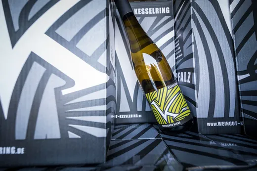 Eine Weinflasche mit einem modernen, geometrischen Etikett lehnt an schwarz-weiß gemusterten Kartons mit den Aufschriften „KESSELRING“ und „WEINGUT-KESSELRING.DE“. Das Design der Kartons passt zum Flaschenetikett.
