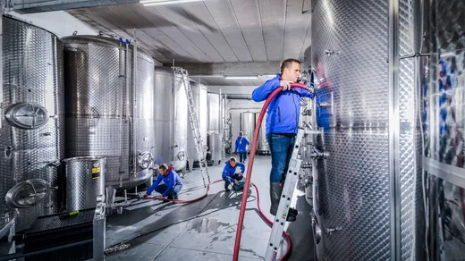 Vier Arbeiter in blauen Uniformen bedienen und überwachen große Edelstahltanks in einer modernen Industrieanlage. Ein Mann steht auf einer Leiter und hantiert mit Schläuchen, die an einem Tank befestigt sind, während andere am Boden mit Geräten arbeiten.