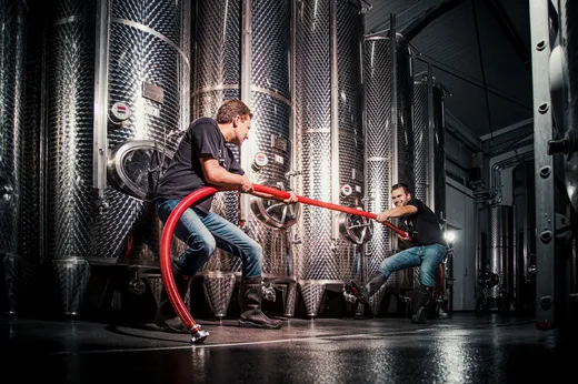 Zwei Personen in einer Weinkellerei oder Brauerei ziehen an den entgegengesetzten Enden eines dicken roten Schlauchs, der zwischen großen Edelstahltanks verläuft; sie tragen schwarze Hemden und Gummistiefel auf dem nassen Boden.