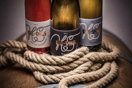 Drei Weinflaschen mit „Kohl-Spiess“-Etiketten – zwei Rotwein- und eine Weißwein-Flasche – stehen auf einem Holzfass, das von einem dicken, gewundenen Seil umschlungen ist.