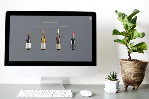 Auf einem Computermonitor ist eine Website mit vier Weinflaschen zu sehen. Der Bildschirm steht auf einem weißen Schreibtisch neben einer Fiddle-Leaf-Feige im Topf und einer kleinen Sukkulente. Der Hintergrund ist hell und minimalistisch gehalten.