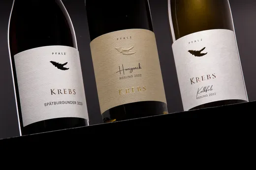 Drei Weinflaschen von Krebs Pfalz: Spätburgunder 2021 (rot), Honigsack Riesling 2022 (beiges Etikett) und Kalkfels Riesling 2022 (weißes Etikett), jeweils mit einem Logo in Form einer Krabbenklaue.