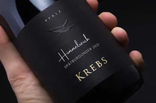 Eine Hand hält eine dunkle Weinflasche mit einem schwarzen Etikett, auf dem in goldener Schrift „PFALZ“, „Himmelreich Spätburgunder 2021“ und „KREBS“ steht. Auf dem Etikett ist außerdem ein kleines Logo in Form einer Krebsklaue zu sehen.
