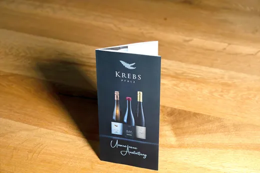 Eine schwarze Weinbroschüre mit dem Titel „Krebs Pfalz“ steht aufrecht auf einer Holzunterlage; auf dem Titelblatt sind Abbildungen von drei verschiedenen Weinflaschen zu sehen.