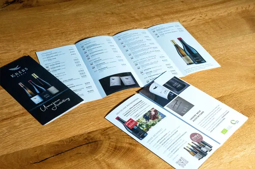 Auf einem Holztisch liegen drei Weinprospekte, die Abbildungen von Weinflaschen, Beschreibungen und Preise zeigen. Die Prospekte sind teilweise aufgeschlagen, sodass verschiedene Weinsorten und Werbeinformationen zu sehen sind.