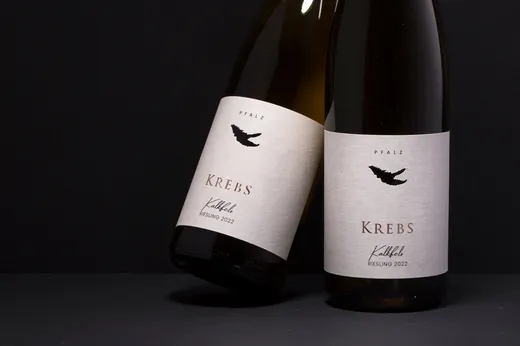 Zwei Weinflaschen mit der Aufschrift „Krebs Riesling 2022 Pfalz“ und minimalistischen weißen Etiketten, auf denen ein Krebsmotiv zu sehen ist. Eine Flasche steht aufrecht, die andere liegt auf der Seite.