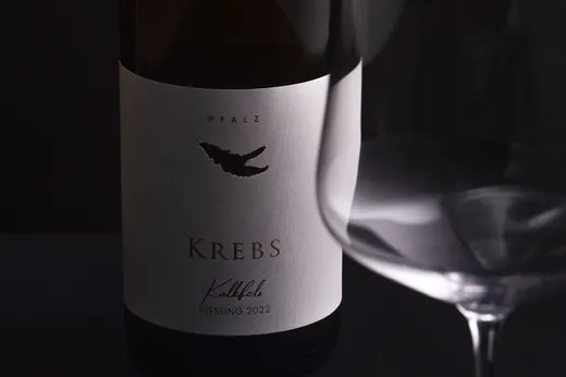 Eine Nahaufnahme einer Weinflasche mit der Aufschrift „Krebs Kalkfels Riesling 2022“ aus der Pfalz steht neben einem leeren Weinglas vor einem dunklen Hintergrund. Auf dem Etikett ist eine schwarze Illustration einer Krabbenklaue zu sehen.