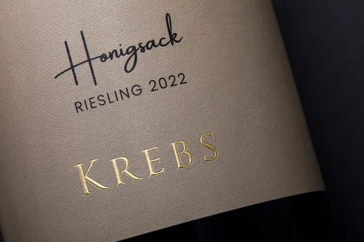 Nahaufnahme eines Weinetiketts mit der Aufschrift „Honigsack“ in schwarzer Schrift und darunter „RIESLING 2022“. „KREBS“ ist in Goldfolie gedruckt.