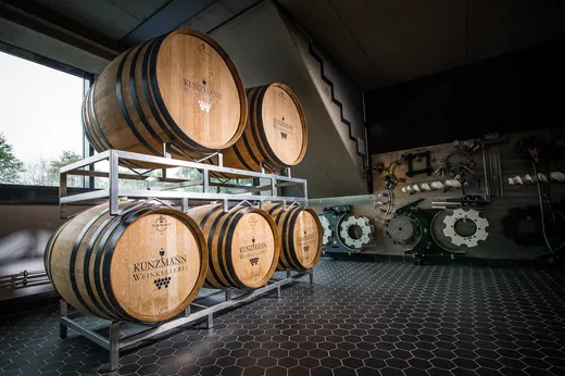 Vier große Holzweinfässer mit der Aufschrift „Kunzmann Weinkellerei“ stehen auf einem Metallgestell in einem modernen Raum mit sechseckigen Bodenfliesen, Betonwänden und Industrieanlagen. Licht fällt durch ein Fenster auf der linken Seite herein.