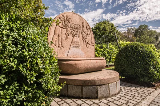 Eine Gedenkbrunnen aus Stein mit eingemeißelten Figuren und der Inschrift „Albertus Quelle Augusta“, umgeben von grünen Sträuchern und Bäumen unter einem teilweise bewölkten Himmel.