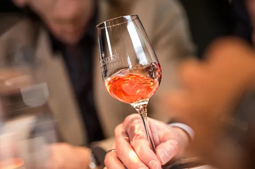 Eine Person hält ein mit Roséwein gefülltes Weinglas in der Hand und schwenkt den Wein darin. Der Hintergrund ist unscharf, der Fokus liegt auf dem Glas und der Hand; im Hintergrund ist eine Person in einer beigen Jacke zu sehen.