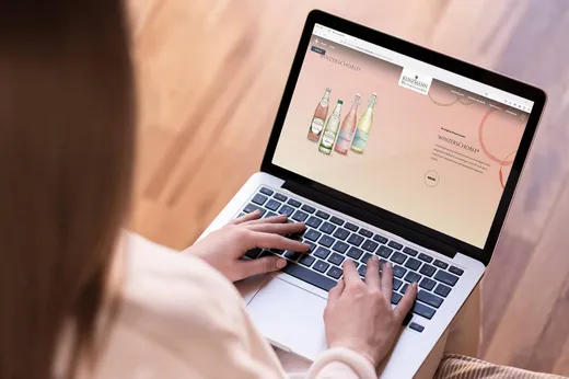 Eine Person, die einen Laptop auf dem Schoß benutzt und eine Website betrachtet, auf der mehrere Flaschen mit kohlensäurehaltigen Getränken vor einem pfirsichfarbenen Hintergrund zu sehen sind.