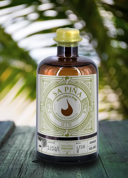 Auf einer grünen Holzunterlage steht eine braune Glasflasche „Sapina Mallorca Pine Gin“ mit einem dekorativen Etikett. Im Hintergrund sind Palmenblätter und strahlendes Sonnenlicht verschwommen zu sehen, was dem Bild eine tropische Atmosphäre verleiht.