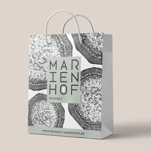 Eine Einkaufstüte aus Papier mit Kordelgriffen ist mit abstrakten schwarz-weißen kreisförmigen Mustern und einem grünen Quadrat mit der Aufschrift „MARIENHOF WEINGUT“ verziert. Unten ist die Website „www.weingut-marienhof.de“ aufgedruckt.