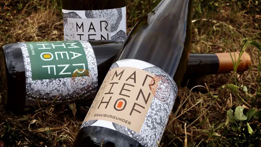 Drei Marienhof-Weinflaschen, deren Etiketten mit moderner, markanter Schrift und strukturierten Motiven versehen sind, liegen im Freien auf Gras und Erde. Zwei Flaschen liegen horizontal, während eine im Hintergrund aufrecht steht.