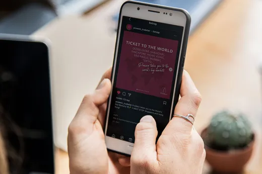 Eine Person, die ein Smartphone in der Hand hält, auf dessen Bildschirm ein Instagram-Beitrag mit Reisezielen zu sehen ist. Im Hintergrund sind ein kleiner Kaktus im Topf und ein Laptop unscharf zu erkennen.