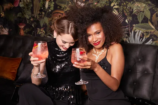 Zwei Frauen in schwarzen Outfits sitzen eng beieinander auf einem Samtsofa, lächeln und halten rosa Cocktails mit Orangenscheiben in der Hand – in einem stilvollen, schwach beleuchteten Raum mit einer Wand mit Blumenmuster.