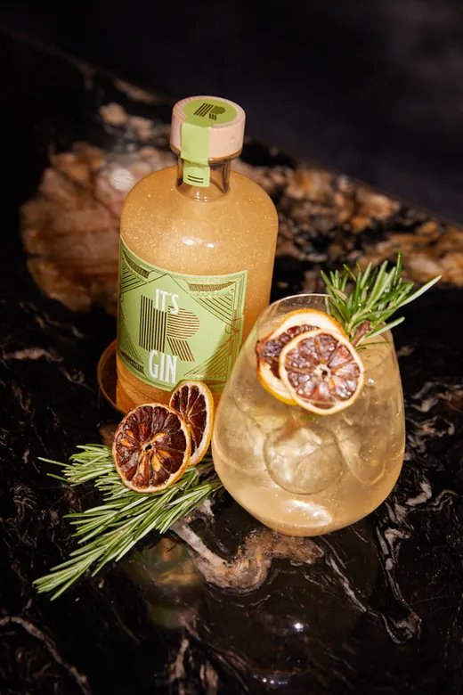 Auf einer dunklen, marmorierten Fläche steht eine Flasche mit der Aufschrift „It's Gin“ neben einem mit Eis gefüllten Cocktailglas, das mit Rosmarin und getrockneten Zitrusscheiben garniert ist.