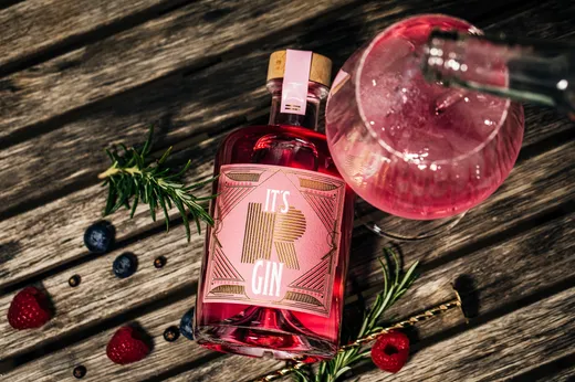 Eine rosa Flasche mit der Aufschrift „It's R Gin“ steht auf einer Holzunterlage neben einem Gin-Cocktail, frischem Rosmarin, Himbeeren, Heidelbeeren und Pfefferkörnern. Aus der Flasche fließt Flüssigkeit in das Glas, wodurch eine festliche Getränkeszene entsteht.