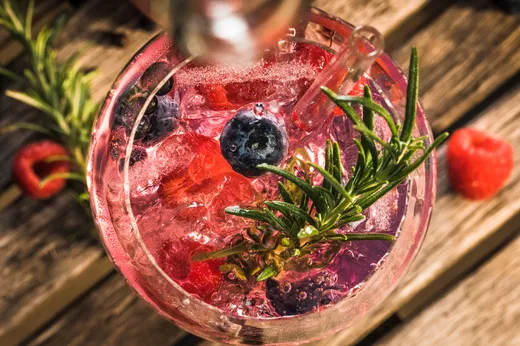 Eine Nahaufnahme eines rosa Cocktails mit Eiswürfeln, Heidelbeeren, Himbeeren und einem Rosmarinzweig in einem Glas auf einer Holzunterlage. Daneben liegen zwei Himbeeren und ein Rosmarinzweig.