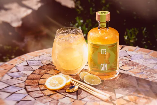 Auf einem Mosaiktisch stehen ein Glas mit einem eisgekühlten, goldfarbenen Gin-Cocktail und eine Flasche mit der Aufschrift „It’s Gin“, daneben liegen Zitronenscheiben, ein Metalllöffel und zwei Cocktailstrohhalme. Sonnenlicht taucht die lebhafte, sommerliche Szene in ein warmes Licht.