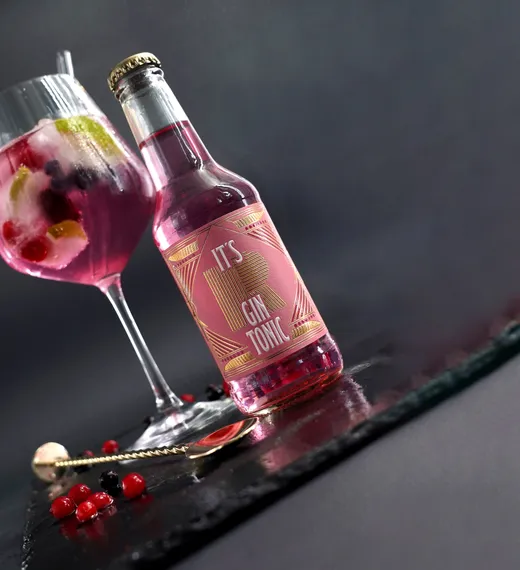 Ein Glas rosa Gin Tonic mit Eis, Limette und Beeren steht neben einer passenden rosa Flasche mit der Aufschrift „IT'S GIN TONIC“ auf einer dunklen Unterlage, auf der Beeren verstreut liegen und ein goldener Löffel liegt.