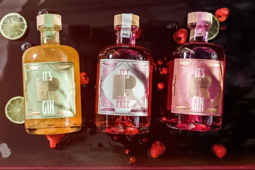 Drei Flaschen „It's R Gin“ in verschiedenen Farben (gelb, rot und rosa) sind auf einer dunklen, nassen Oberfläche zu sehen, umgeben von Limettenscheiben und verstreuten Beeren.