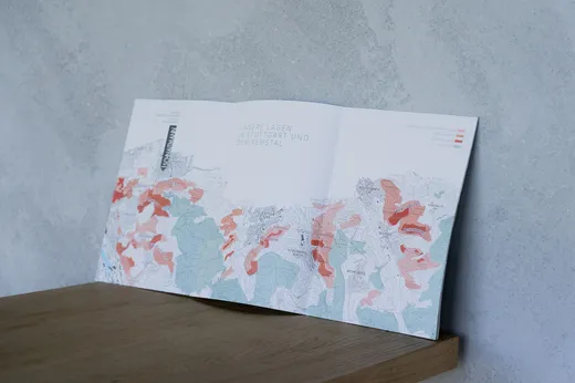 Eine aufgeschlagene Landkarte mit rot, grün und blau schattierten Bereichen, auf der verschiedene Regionen mit Beschriftungen zu sehen sind, liegt auf einer Holzfläche vor einer hellgrauen, strukturierten Wand.