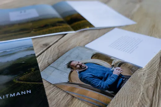 Auf einer Holzfläche liegt eine aufgeschlagene Broschüre, auf der ein Foto eines Mannes in einem blauen Pullover zu sehen ist, der neben großen Holzfässern steht; auf den angrenzenden Seiten sind verschwommene Texte und Bilder zu erkennen.