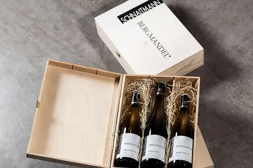 Drei Flaschen Bergmandel-Wein in einer hölzernen Geschenkbox mit Strohpolsterung, auf einer grauen Unterlage platziert. Der mit „SCHNAITMANN“ und „BERGMANDEL“ beschriftete Deckel der Box ist geöffnet und liegt hinter der Box.