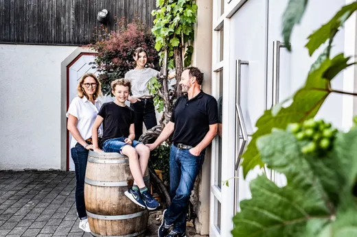 Vier Personen stehen und sitzen im Freien in der Nähe eines Gebäudes um ein Weinfass herum. Sie sind leger gekleidet, lächeln und wirken entspannt. An der Wand ranken grüne Weinreben empor, was eine lebhafte, einladende Atmosphäre schafft.