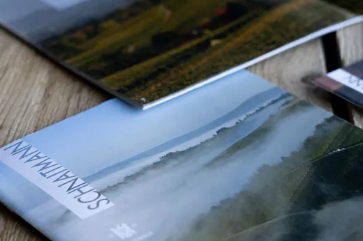 Eine Nahaufnahme von zwei Zeitschriften auf einer Holzfläche; auf dem Cover der einen ist eine neblige Landschaft mit dem Wort „SCHNAITMANN“ zu sehen. Die andere Zeitschrift zeigt eine Luftaufnahme von grünen Feldern.