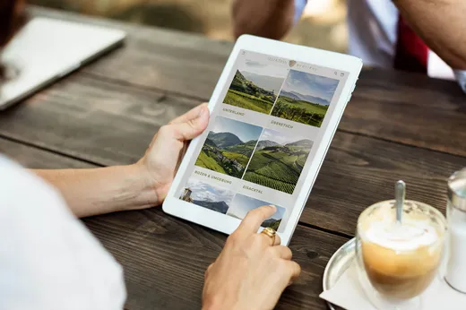 Eine Person, die ein Tablet in der Hand hält und an einem Holztisch im Freien Landschaftsfotos ansieht; daneben stehen ein Cappuccino und eine Untertasse, im Hintergrund ist eine weitere Person teilweise zu sehen.