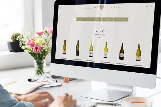 Eine Person sitzt vor einem Desktop-Computer, auf dem eine Website mit einer Weinauswahl angezeigt wird, die verschiedene Flaschen Weißwein zeigt. Auf dem hellen, aufgeräumten Schreibtisch stehen eine Vase mit Blumen und Büromaterial.