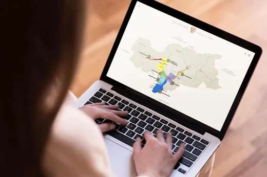 Eine Person benutzt einen Laptop, auf dem eine Karte mit farbigen Gebieten und beschrifteten Punkten zu sehen ist. Die Karte trägt den Titel „SELEKTION SÜDTIROL“ und zeigt geografische Gebiete mit verschiedenen Markierungen. Nur die Hände der Person sind zu sehen.