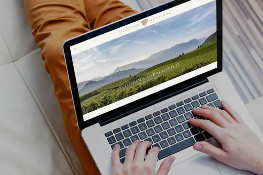 Eine Person sitzt auf einem Sofa und nutzt einen Laptop, auf dem eine Website mit einem Landschaftsfoto und einem deutschen Text mit der Überschrift „Südtirols Anbaugebiete“ unter einem klaren Himmel zu sehen ist.